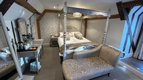 Prop-Search A bedroom in a hotel, showing a room decorated mostly in grey and silver, showing a bed, small chaise longue, a desk, with items on it, a four-poster bed, window to the right, door to the left and mirrors. 