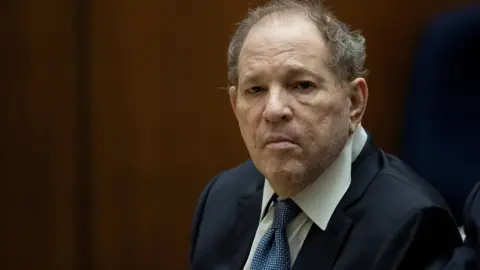 Reuters  Former film producer Harvey Weinstein appears in court. He is wearing a blue suit and a white shirt with a rumpled collar. His tie is blue.