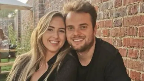 Health and Safety Executive A man with dark hair and a woman with blonde hair smile at the camera with a brick wall behind them