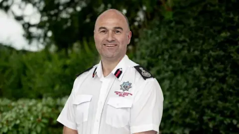 A headshot of a man standing in a garden, wearing a white shirt with the fire service logo on the right. He is smiling at the camera. 