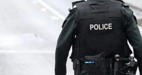 A police officer is standing on a road. They are in a black uniform and have a belt with a number of items. 