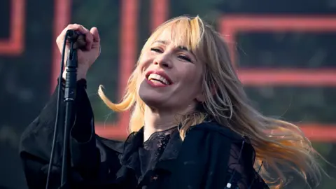 Natasha Bedingfield wearing a black top performing on stage. She is smiling and holding a microphone. She has blonde hair and is wearing a black top. 