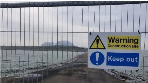 A sign warning of a construction site fixed to a wire fence in front of frosty grass and a small road leading to two domes.