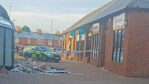 Laura Barford A police car, in a shopping area, with debris on the ground to the left. There are shops to the right and houses behind the police car. There is police tape across the front of the buildings. Several people are standing about in the distance. 