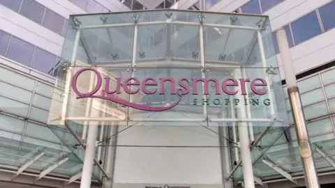 LDRS A glass sign above a set of double doors saying "Queensmere shopping" in pink and green writing.