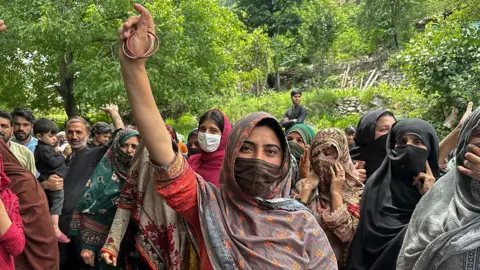 A crowd of protesters, mainly women, gathers, with some raising their hands. their faces are covered with headscarves.