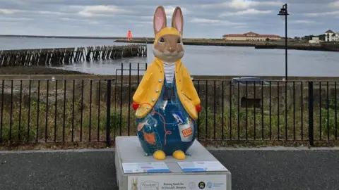 A large Peter Rabbit sculpture dressed in a yellow fisherman's hat and jacket standing in front of the sea. Underneath it is wearing blue dungarees with various fishing equipment painted on. There is a black fence behind it with some shrubbery behind it, before the sea. There are buildings on the right and a small red lighthouse immediately behind it on a pier.