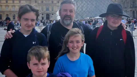 Jonathan Sivyer is standing in the middle of his four children in front of the iconic Louvre Museum pyramid.