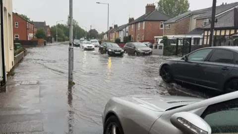 A road is flooded and several cars are driving through it on either side of the road. The water has come up on to the pavement is going near people's houses either side of the road. 