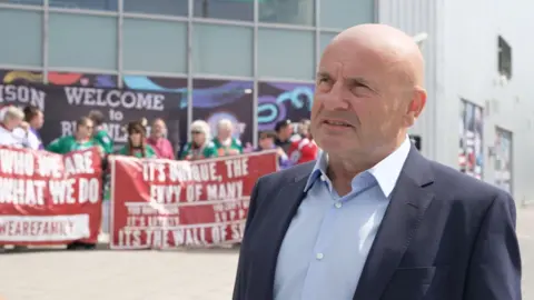 Steve Nell stands outside Planet Ice Basingstoke surrounded by fans holding red banners supporting the Hampshire based team