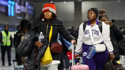 Two Ghanaian women in winter outfits, one in a Boston sweatshirt, wheeling suitcases at OR Tambo Airport in Johannesburg - 27 May 2026
