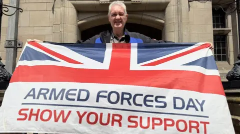 A man is standing outside a large stone building, holding a flag. It has half a union jack on it, with the words "armed forces day, show your support" on the bottom half.