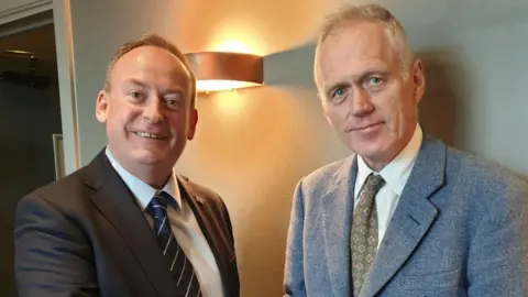 Deputy Lyndon Trott can be seen pictured on the left wearing a dark grey suit, white shirt and a navy blue tie with diagonal white pin stripes. He has short mousy brown hair. Lord Ponsonby is pictured on the right wearing a light blue wool suit. He is wearing a white shirt with a green tie with diamond patterning. He has white, thin hair. Both men are pictured in a room with olive coloured walls with a light behind them.