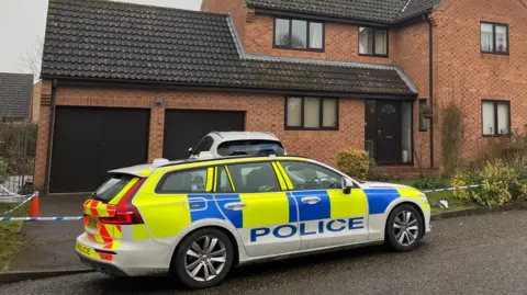 Ant Saddington/BBC A police car is outside a property in Lingwood Park in Peterborough. Around the house is blue and white tape as a police cordon. 