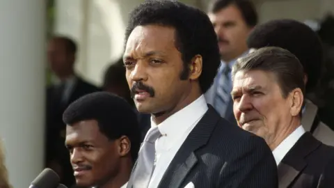 Civil rights activist Jesse Jackson speaks during a ceremony in honor of Navy pilot Robert O. Goodman with President Ronald Reagan stood behind him in 1984