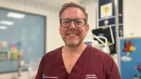 Consultant neonatologist Matt Nash is opinionated successful a infirmary area. He is wearing maroon scrubs pinch his sanction connected his top. He has a beard and is wearing glasses. There is infirmary instrumentality down him and a brightly coloured picture.
