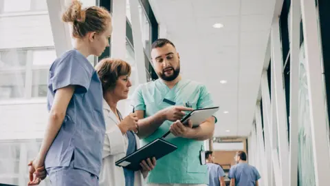 Getty Images Male healthcare worker explaining over clipboard to doctor at hospital - stock photo. he wears green scrubs, has a beard and short hair. The two people he is talking to are female medics. The one on the left is in blue scrubs and has curly blonde hair tied up. The one in the middle wears a white coat and has short hair.