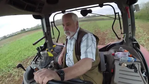 BBC A man sat at the wheel of a tractor with the fields seen behind and to the side from the inside of his tractor.