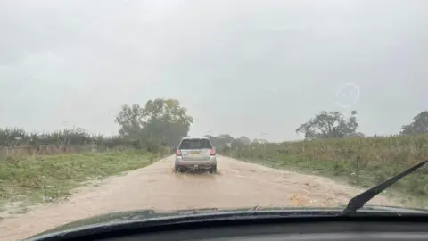 Kate Fraser A wide view of a road from inside a car. There is a car driving through brown muddy floodwater which covers a road.