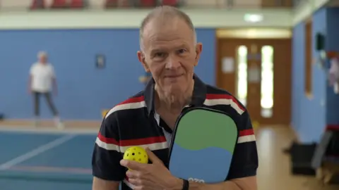 Jamie Niblock/BBC Jonathan Burney standing on an indoor pickleball court at Hingham Sports Centre in Norfolk. He is looking directly at the camera and smiling. He is wearing a white, red and black striped polo shirt and is holding a pickleball racket under his arm and holding a yellow ball in his hand.