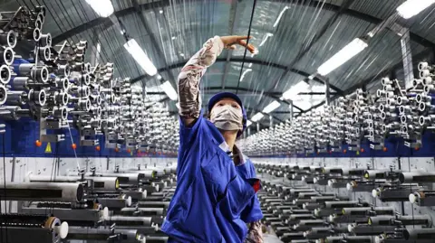 In a factory, a woman in a blue uniform and face mask reaches up with her right hand to hold a string above her head