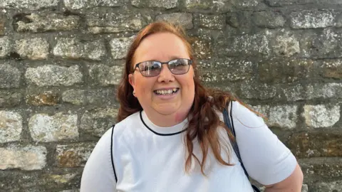 Red haired woman, wearing glasses and a white short sleeved knit top smiling while looking at the camera. She is standing in front of a stone wall.