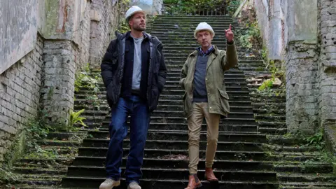 Sky Edwards and Baker walk side-by-side down stairs at the castle. Both are wearing hard hats, with Baker pointing to the sky