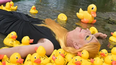 Mark Coller A man in drag wears a black formal dress and an orange wig. He is lying down in water and surrounded by rubber ducks with numbers on them.