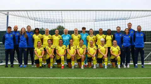 Lucy Copsey A football team photo. The players and staff are standing or crouching down in front of a large goal net. The players are wearing a yellow kit and the staff are wearing a blue kit.