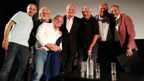 Jimmy Daly A group of smartly dressed middle aged men huddle together with their arms around each other on a stage