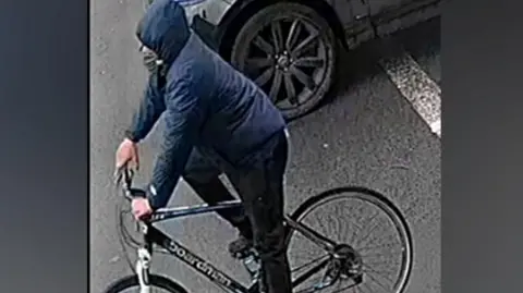 A man on a bike against a grey background. He is wearing a blue coat with the hood up and black trousers and is on a black bike with a white bar by the wheel. 