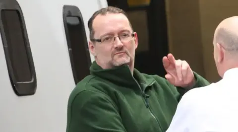 Tim Bugler A man with glasses and a green top makes a gesture to the camera as he is led from a prison van. 