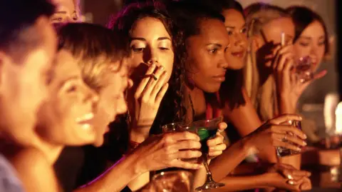 Getty Images Nine people, mostly women, in a line at a bar, smoking cigarettes and holding alcoholic drinks. They all look dressed up for a fun night out.  