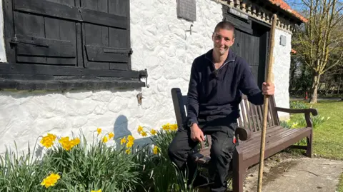 Tom Harvey is sitting on the arm of a brown wooden bench while outside. In front of him is a patch of grass with daffodils. He is wearing black cargo trousers, a blue 3/4 zip jumper and has a long wooden pole in his left hand. He is smiling at the camera and has short dark hair.