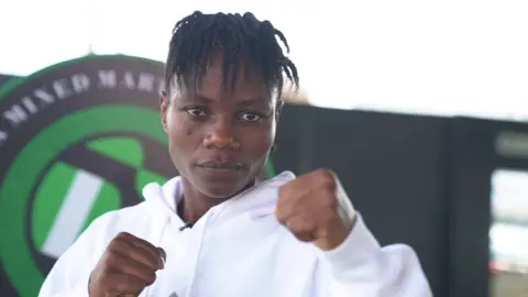 Juliet Ukah wears a white hoodie, looks into the camera and holds up her hands in a fighter's pose with a green and black logo on the wall behind her
