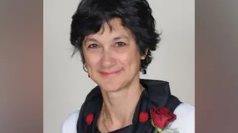 A grainy image showing Katie Thornburrow who has short dark brown hair and is wearing a white top with a black cowl collar, with a red rose pinned to her left shoulder. She is smiling.
