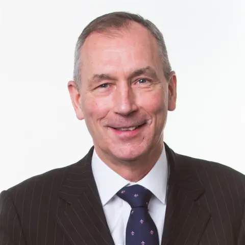 Caeli Studio-style head-and-shoulders portrait of a man wearing a dark pinstriped suit, white shirt, and patterned tie against a plain light-grey background. The individual faces the camera directly, with the upper body centred in the frame.
