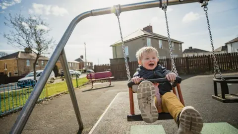 Getty Images A young child is sitting on a swing. He is in a playground with houses in the background and a blue sky above.