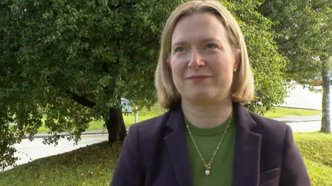 A woman in a navy suit jacket, green knit top under with gold pendant necklace looks at the camera with a close mouthed smile. She has a short blond bob and is standing in front of several large trees.