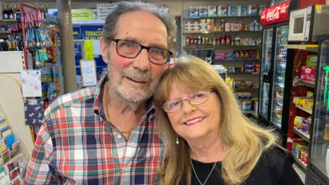 A man and woman lean on each other while they smile at the camera. They are standing in a convenience store with produce on shelves in the background.