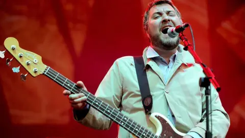 Gambar Getty Jimi Goodwin, dengan rambut pendek dan janggut, mengenakan jaket krim pucat dan kemeja putih saat ia memainkan gitar dan bernyanyi ke dalam mikrofon di depan latar belakang merah