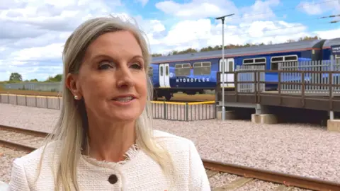A woman with blonde hair is wearing a white coat and standing in front of the railway test track. A train and platform are visible behind her.
