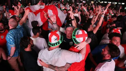 Getty Three England fans in St George's Cross bucket hats embrace amid a packed crowd at Boxpark Croydon. A large England flag is held aloft behind them.