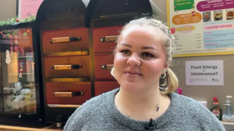 A young woman with blonde hair scraped back into a pigtail smiles at the camera. She is standing in front of a large oven containing jacket potatoes.