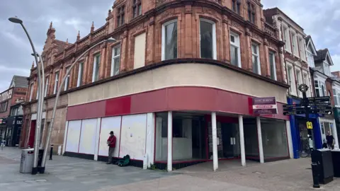 A building on a corner of a street has a shop empty on its first floor. Above it are two more storeys which have large windows. A person is playing a guitar on the street outside of the building.