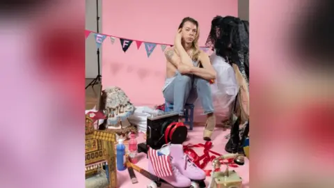 Paul Blakemore Tom Marshman wearing denim jean in a room with items symbolic of him. They include pink roller boots, a US flag, a lampshade, paints, net outfits and a boom box, all with a pink background.