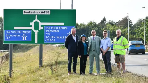 A group of five councillors standing on a grassy verge beside the road. There is a large sign behind them signalling the upcoming roundabout exits for the South West (Gloucester) M5, Tewkesbury A438, and The Midlands and South Wales.