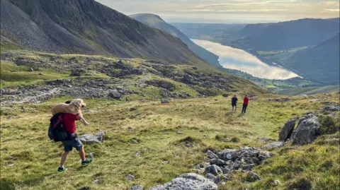 A rescuer in a red t-shirt carries a large golden retriever dog across his shoulders while descending Scafell Pike in the Lake District, In the background is a mountainous landscape and Wastwater, a body of water at the foot of the mountain.