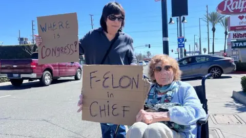 BBC One woman stands and holds sign that reads "Where is congress", a second one woman next to her sits in wheelchair and holds sign that reads "F-Elon in Chief" while cars drive on a busy street in the background