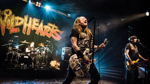 Getty Images Vocalist and guitarist Ginger Wildheart (L) and bassist Danny McCormack of English hard rock group The Wildhearts performing live on stage at KOKO in London, on 2 December 2018. Wildheart has a low slung white semi-acoustic six string guitar with many images on it, reminiscent of tattoo flash art. He is wearing a black t-shirt with an image on the front. McCormack has a mustard and black bass and wears a black peaked cap. Behind them is the drummer behind a black kit and the Wildhearts logo, in orange and yellow, is hanging on a black fabric behind the drum riser.